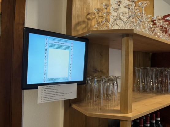 Ein Bildschirm mit Hinweisen zur Abrechnung von Getränken ist an einer Wand neben einem Holzregal mit Reihen von Gläsern und Weinflaschen angebracht.