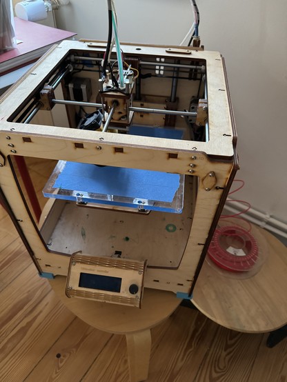 Ein 3D Drucker aus Holz. Daneben eine Spule rotes Filament. 