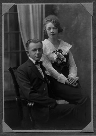 Schwarz-Weiß-Foto eines sitzenden Paares, der Mann im Anzug mit Fliege und die Frau im Kleid mit Korsage, beide mit Blick in die Kamera.