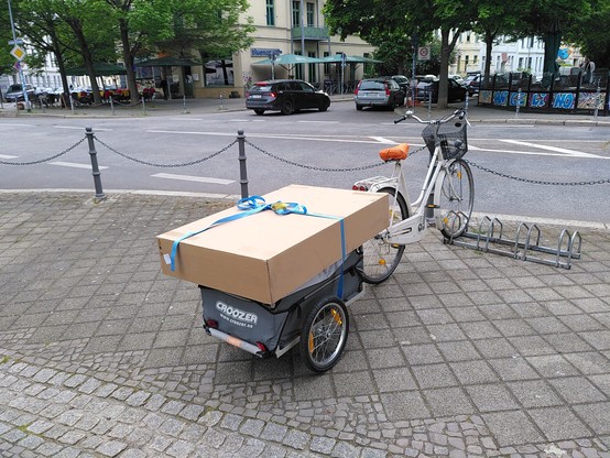 Fahrrad mit Anhänger, auf dem ein ca. 110cm x 70cm großes Paket liegt.