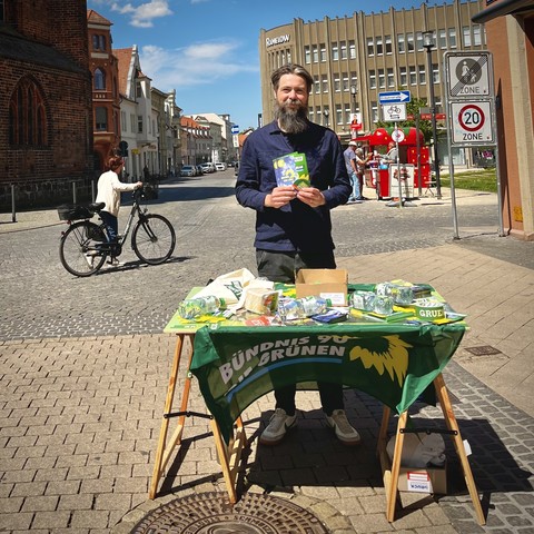 A bearded man standing at a foldable table with pamphlets and promotional materials for the Green Party on a sunny pedestrian street. Behind him, there is a cyclist and various city buildings.