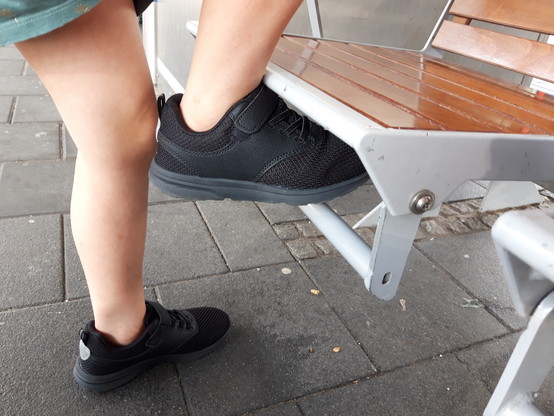 Ein Wartesitz aus Holz und Metall auf einem Bahngleis. Eine Person steht davor und hat ein Bein zwischen Sitzfläche und darunter liegender freischwingender Metallstange gesteckt.