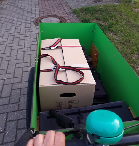 In einer grünen Lastenradkiste steht ein Umzugskarton, der mit Spanngurten verschlossen ist.