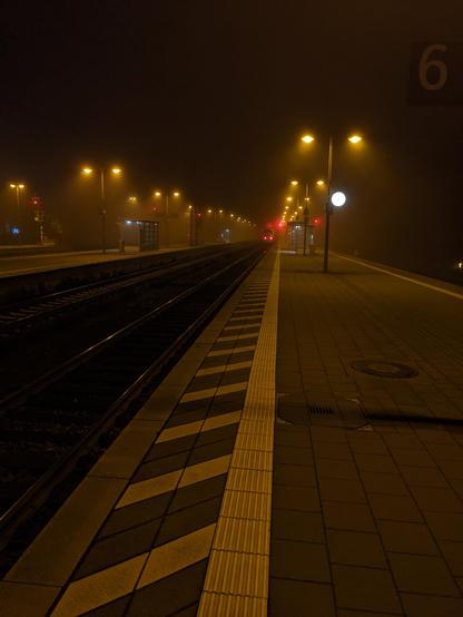 Eine Bahnsteigkante. Am Ende sind noch die Schlusslichter eines ICE neben einem Signal zu erahnen (rot). Es ist Abend und die Beleuchtung des Bahnsteigs ist an, das Licht scheint diffus im Nebel.