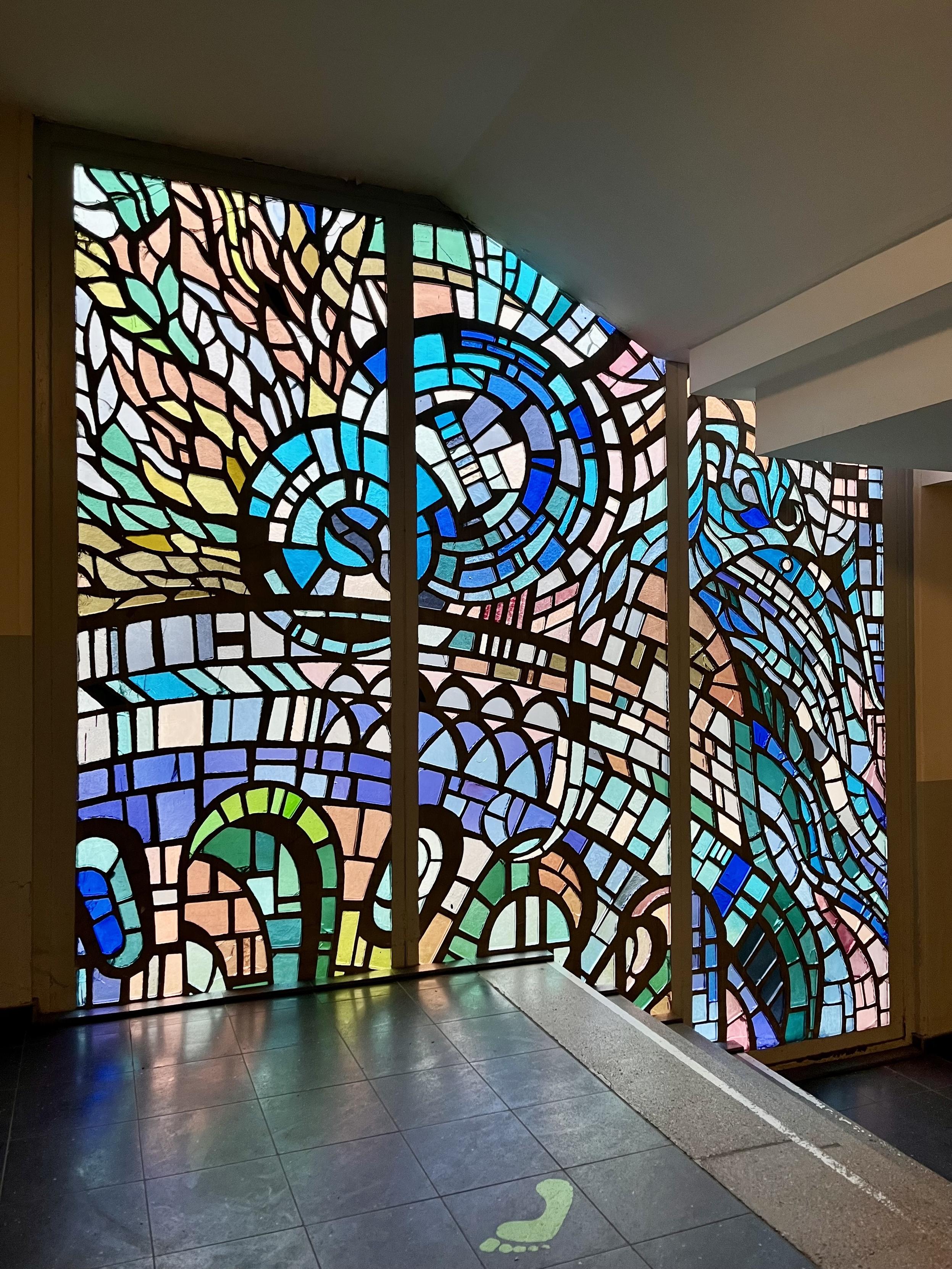 Farbenfrohe Buntglasfenster mit filigranen geometrischen Mustern in Blau-, Grün- und Pinktönen sowie klarem Glas, installiert in einem modernen Ambiente. Auf dem Boden darunter ist ein grüßer Fußabdruck zu sehen.